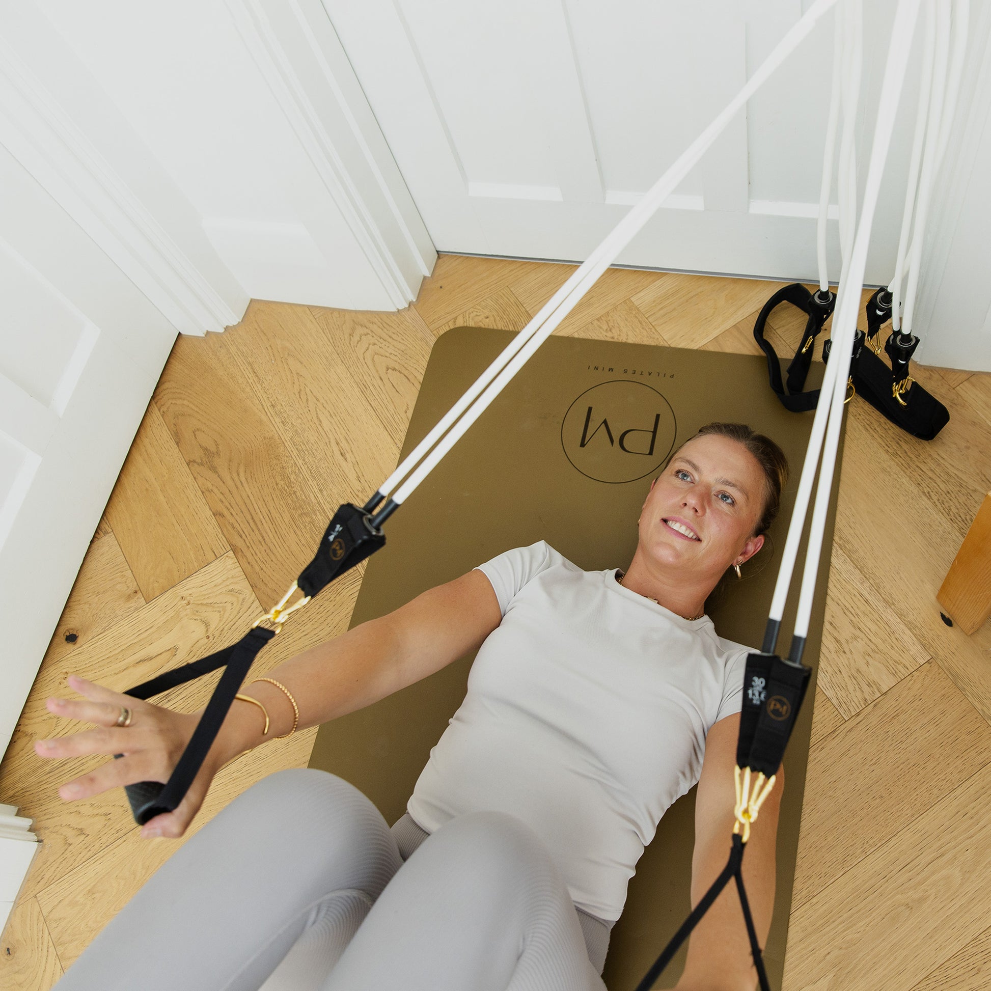 A woman lies on a brown mat on a light wood floor, using a Resistance bands of Pilates Mini Kit trainer with white straps and black handles.
