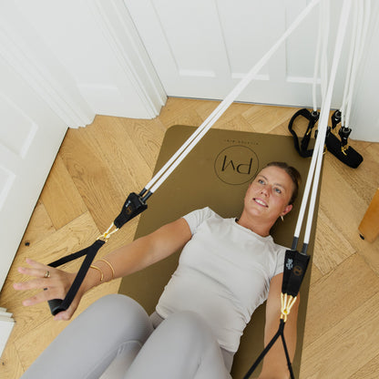 A woman lies on a brown mat on a light wood floor, using a Resistance bands of Pilates Mini Kit trainer with white straps and black handles.