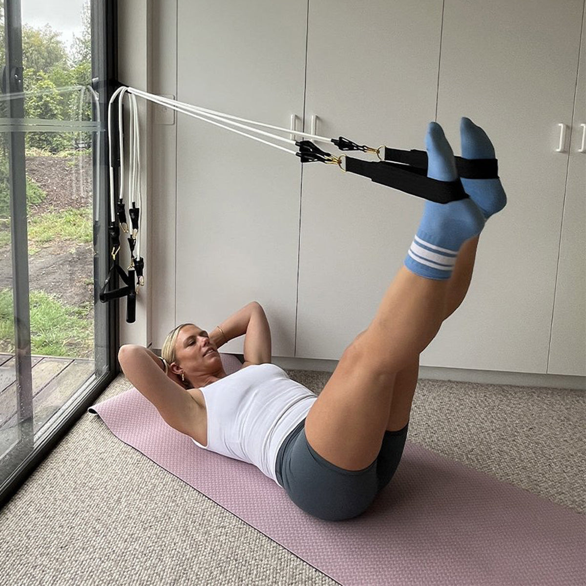 A woman lies on a pink exercise mat in a bright room, using a Pilates Mini resistance band with ankle straps attached to a window-mounted frame. She wears a white tank top, dark gray shorts, and light blue socks with white stripes.