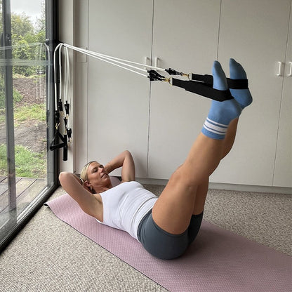 A woman lies on a pink exercise mat in a bright room, using a Pilates Mini resistance band with ankle straps attached to a window-mounted frame. She wears a white tank top, dark gray shorts, and light blue socks with white stripes.